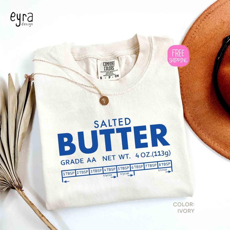 May include: Ivory-colored t-shirt with the words "SALTED BUTTER" in blue, along with measurement conversions. The shirt also features the text "GRADE AA NET WT. 4 OZ. (113g)". A gold necklace with a pendant is draped on the shirt.