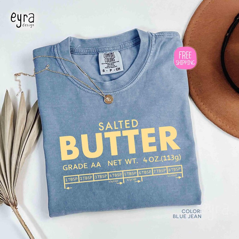 May include: A blue jean colored t-shirt with the words "SALTED BUTTER" in yellow block letters. The shirt also features a graphic with measurements and the text "GRADE AA NET WT. 4 OZ. (113g)". A gold necklace rests on the shirt.