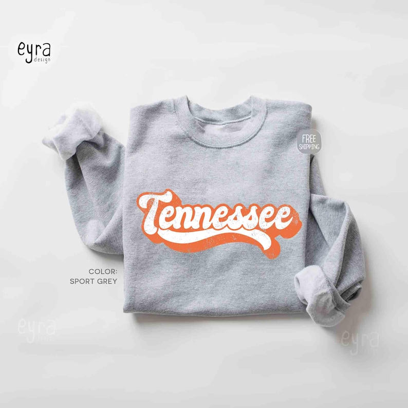 K&ouml;nnte beinhalten: Ein sportgraues Sweatshirt mit Rundhalsausschnitt und dem Wort Tennessee, das in einer kr&auml;ftigen, orangefarbenen, kursiven Retro-Schrift auf der Brust gedruckt ist. Das Kleidungsst&uuml;ck hat lange &Auml;rmel mit gerippten B&uuml;ndchen und einen gerippten Ausschnitt.