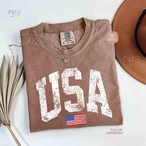 Puede incluir: Camiseta color espresso con la palabra "USA" en letras blancas desgastadas y una peque&ntilde;a bandera americana. Un collar plateado con un colgante descansa sobre la camiseta. La camiseta es de la marca Comfort Colors.