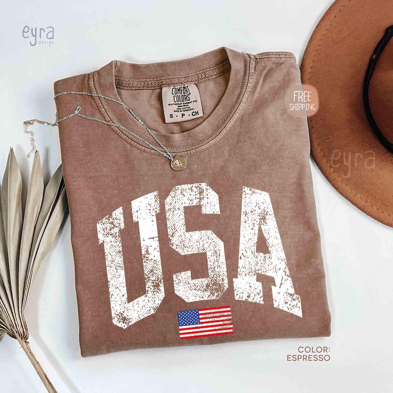 Puede incluir: Camiseta color espresso con la palabra "USA" en letras blancas desgastadas y una peque&ntilde;a bandera americana. Un collar plateado con un colgante descansa sobre la camiseta. La camiseta es de la marca Comfort Colors.