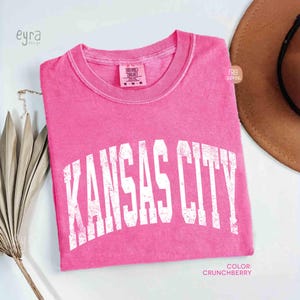 May include: A bright pink t-shirt with the text "KANSAS CITY" in white, arched lettering. The shirt is a solid color and has a crew neck. The color is described as "Crunchberry".