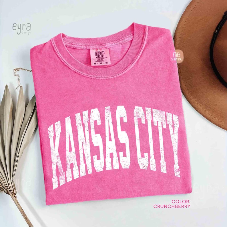 May include: A bright pink t-shirt with the text "KANSAS CITY" in white, arched lettering. The shirt is a solid color and has a crew neck. The color is described as "Crunchberry".