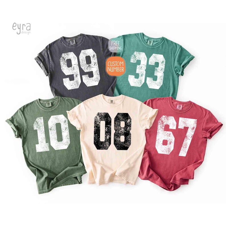 K&ouml;nnte beinhalten: F&uuml;nf kurz&auml;rmelige T-Shirts in verschiedenen Farben, jedes mit einer gro&szlig;en, verwitterten Nummer. Farben sind Anthrazit, Gr&uuml;n, Creme, T&uuml;rkis und Rot. Die Nummern sind wei&szlig; oder schwarz. Die Shirts sind wahrscheinlich f&uuml;r Sport oder Team-Events.