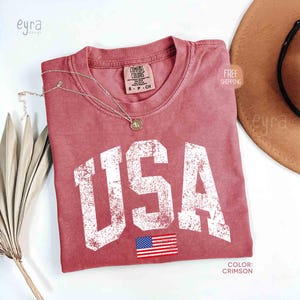 Puede incluir: Camiseta de color carmes&iacute; con la palabra "USA" en letras blancas desgastadas y una peque&ntilde;a bandera americana. Un collar dorado descansa sobre la camiseta. La camiseta est&aacute; etiquetada como "Comfort Colors" y es talla S-P-CH.