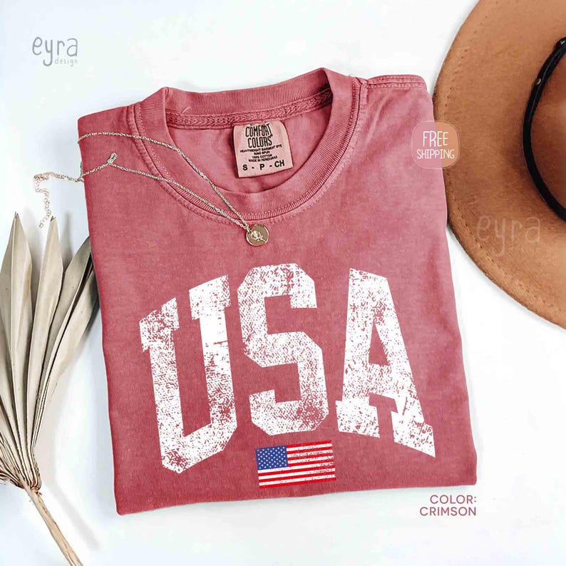 Puede incluir: Camiseta de color carmes&iacute; con la palabra "USA" en letras blancas desgastadas y una peque&ntilde;a bandera americana. Un collar dorado descansa sobre la camiseta. La camiseta est&aacute; etiquetada como "Comfort Colors" y es talla S-P-CH.