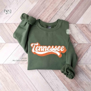 K&ouml;nnte beinhalten: Ein milit&auml;rgr&uuml;ner Rundhals-Pullover mit dem Wort Tennessee, das in einer abgenutzten wei&szlig;en Schrift mit kr&auml;ftiger orangefarbener Umrandung aufgedruckt ist. Das Kleidungsst&uuml;ck hat lange &Auml;rmel mit gerippten B&uuml;ndchen und einen gerippten Ausschnitt.