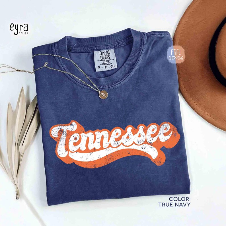 May include: A navy blue t-shirt with the word "Tennessee" in white and orange lettering. The shirt has a crew neck and short sleeves. A gold necklace with a pendant is draped across the shirt.