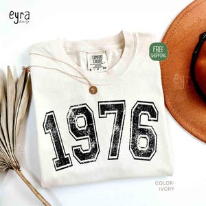 May include: Ivory-colored t-shirt with the year "1976" printed in distressed black lettering. A gold necklace with a pendant rests on the shirt. The shirt is labeled "Comfort Colors" and the color is "Ivory".