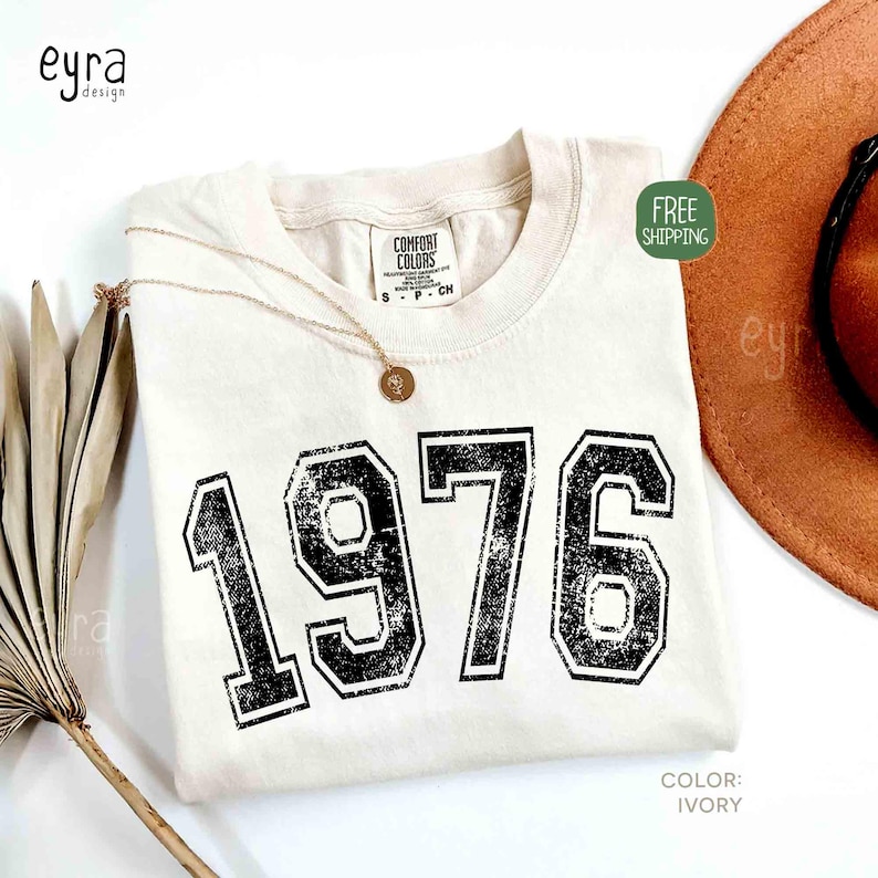 May include: Ivory-colored t-shirt with the year "1976" printed in distressed black lettering. A gold necklace with a pendant rests on the shirt. The shirt is labeled "Comfort Colors" and the color is "Ivory".
