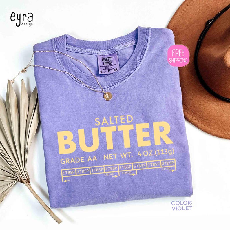 May include: A violet-colored t-shirt with the words "SALTED BUTTER" in yellow block letters. The shirt also features a measurement guide and the text "GRADE AA NET WT. 4 OZ. (113g)." A gold necklace and a brown hat are also visible.