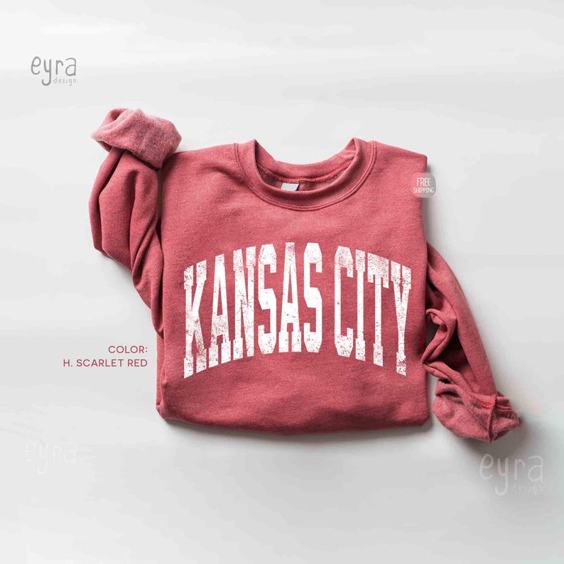 May include: A crimson sweatshirt featuring the white text "KANSAS CITY" in an arched design. The garment has a crew neck and long sleeves with rolled cuffs. The colour is described as H. Scarlet Red.