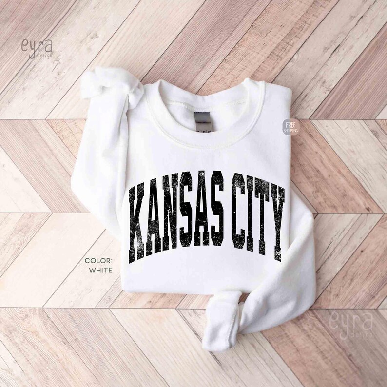 May include: A white sweatshirt with the text "KANSAS CITY" in distressed black lettering. The sweatshirt is folded on a light wood surface. The colour of the sweatshirt is white.