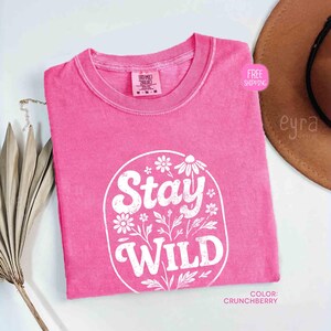 May include: A pink t-shirt with the words "Stay Wild" in a white floral design. The shirt is a solid color and has a crew neck. The color is "Crunchberry".