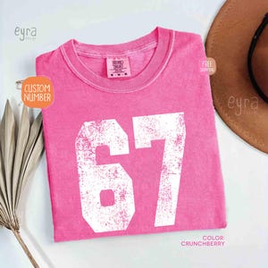 K&ouml;nnte beinhalten: Ein pinkfarbenes T-Shirt mit der Nummer 67 in wei&szlig;er, abgenutzter Schrift. Das Shirt hat einen Rundhalsausschnitt und ein Etikett am Kragen. Die Farbe wird als "Crunchberry" bezeichnet. Die Worte "Custom Number" stehen in einem orangefarbenen Kreis.