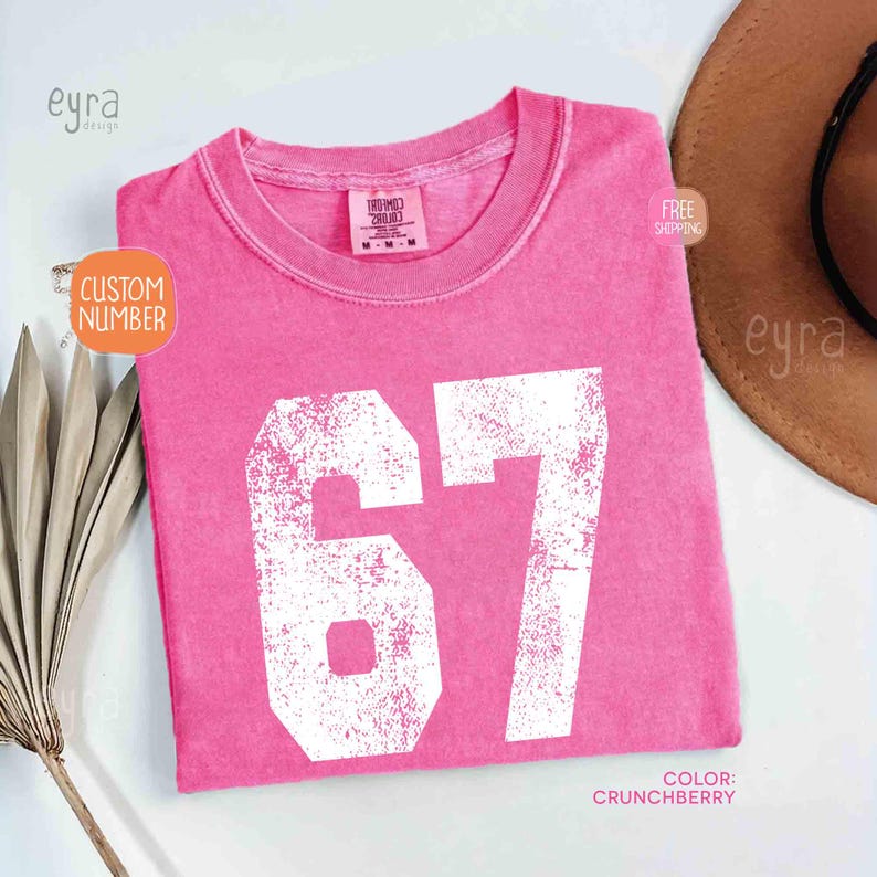 K&ouml;nnte beinhalten: Ein pinkfarbenes T-Shirt mit der Nummer 67 in wei&szlig;er, abgenutzter Schrift. Das Shirt hat einen Rundhalsausschnitt und ein Etikett am Kragen. Die Farbe wird als "Crunchberry" bezeichnet. Die Worte "Custom Number" stehen in einem orangefarbenen Kreis.