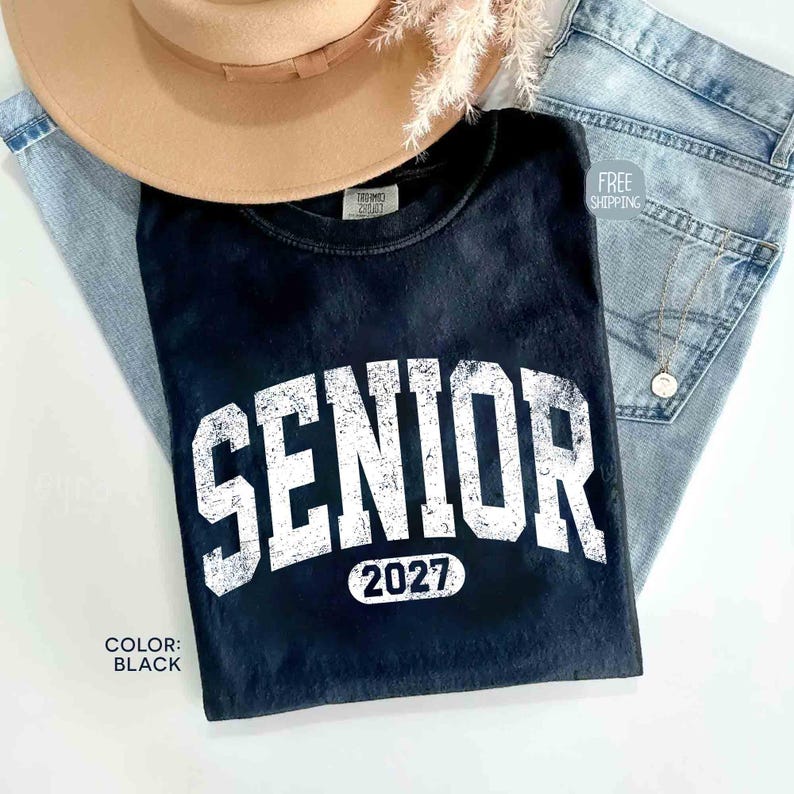 May include: Black t-shirt with the word "SENIOR" in large white block letters and the year "2027" underneath. The shirt is paired with light-wash denim jeans and a tan hat. The shirt's color is black.