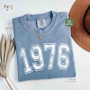 May include: A blue jean colored t-shirt with the number "1976" printed in white. The shirt has a crew neck and short sleeves. A gold necklace with a pendant is draped on the shirt. The shirt is labeled "Comfort Colors".