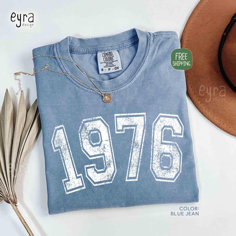 May include: A blue jean colored t-shirt with the number "1976" printed in white. The shirt has a crew neck and short sleeves. A gold necklace with a pendant is draped on the shirt. The shirt is labeled "Comfort Colors".