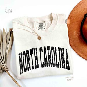 K&ouml;nnte beinhalten: Ein elfenbeinfarbenes Kurzarm-T-Shirt mit Rundhalsausschnitt und einem Comfort Colors Markenetikett. Das Shirt zeigt den Schriftzug NORTH CAROLINA in einer abgenutzten schwarzen Schrift auf der Brust. Eine d&uuml;nne goldene Kette liegt auf dem Stoff.