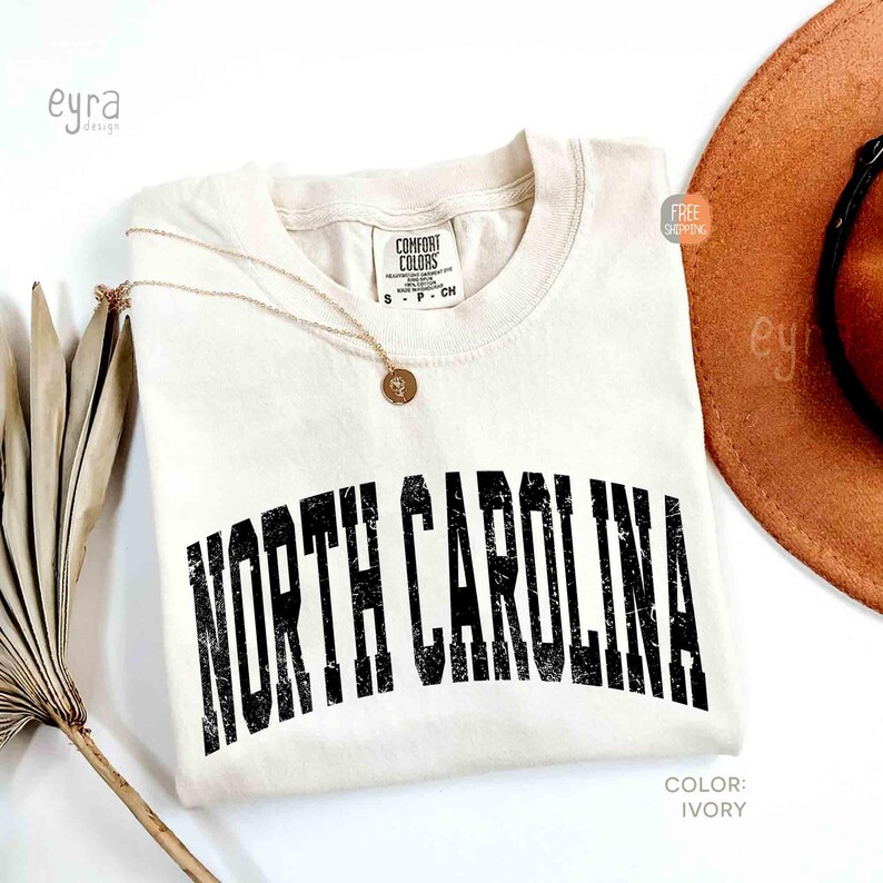 K&ouml;nnte beinhalten: Ein elfenbeinfarbenes Kurzarm-T-Shirt mit Rundhalsausschnitt und einem Comfort Colors Markenetikett. Das Shirt zeigt den Schriftzug NORTH CAROLINA in einer abgenutzten schwarzen Schrift auf der Brust. Eine d&uuml;nne goldene Kette liegt auf dem Stoff.