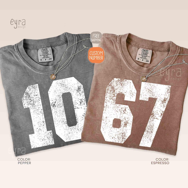 K&ouml;nnte beinhalten: Zwei Comfort Colors T-Shirts, eines in Pfeffergrau und eines in Espressobraun, jeweils mit einer gro&szlig;en, wei&szlig;en, verwitterten Zahl. Das graue Shirt zeigt "10", das braune Shirt zeigt "67". Beide haben einen Rundhalsausschnitt und ein kleines Etikett.