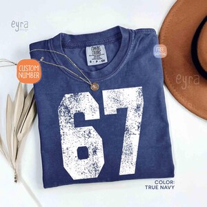 K&ouml;nnte beinhalten: Ein marineblaues T-Shirt mit der Nummer 67 in abgenutzten wei&szlig;en Buchstaben. Das Shirt hat einen Rundhalsausschnitt und ein kleines Etikett. Eine goldene Halskette mit Anh&auml;nger liegt auf dem Shirt. Der Text "CUSTOM NUMBER" steht auf einem Anh&auml;nger.