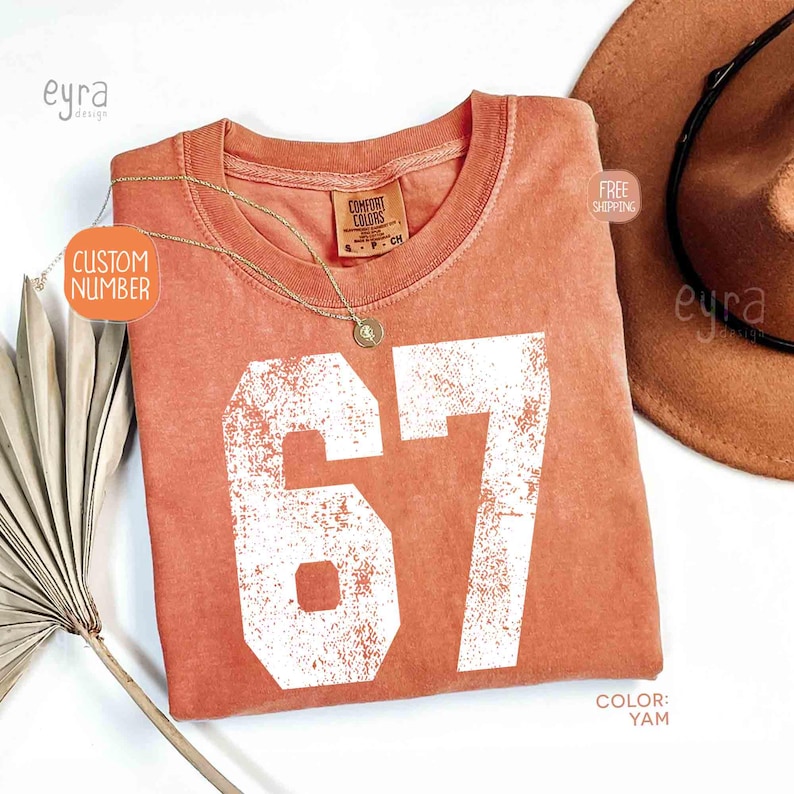 K&ouml;nnte beinhalten: Ein korallenfarbenes T-Shirt mit der Nummer 67 in wei&szlig;er, verblichener Schrift. Eine goldene Halskette mit Anh&auml;nger liegt auf dem Shirt. Ein brauner Hut ist im Hintergrund zu sehen. Das Shirt hat ein Comfort Colors-Etikett.