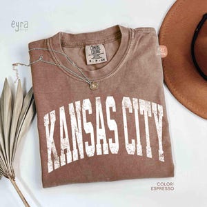 May include: Espresso-colored t-shirt with the text "KANSAS CITY" in white, distressed lettering. The shirt is a Comfort Colors brand, size S-P-CH. A silver necklace with a pendant and a brown hat are also visible.
