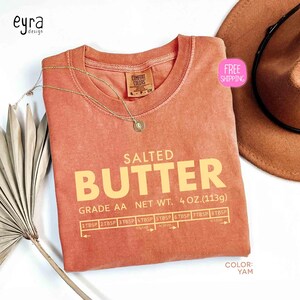 May include: A yam-colored t-shirt with the words "SALTED BUTTER" in large, yellow letters. The shirt also features a butter measurement guide and the text "GRADE AA NET WT. 4 OZ. (113g)." A gold necklace and a brown hat are also visible.
