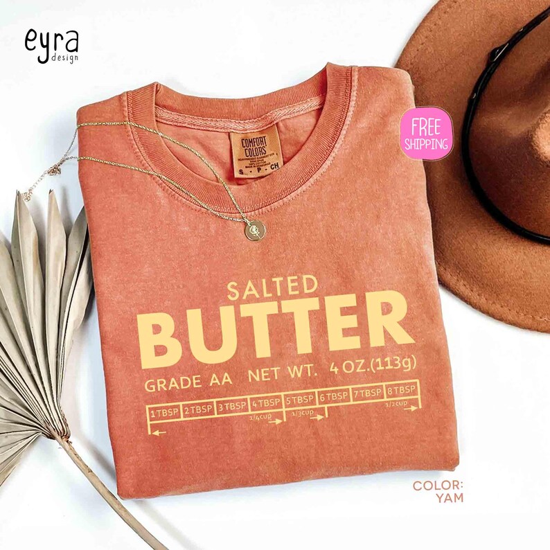 May include: A yam-colored t-shirt with the words "SALTED BUTTER" in large, yellow letters. The shirt also features a butter measurement guide and the text "GRADE AA NET WT. 4 OZ. (113g)." A gold necklace and a brown hat are also visible.