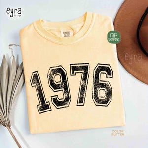 May include: Butter-colored t-shirt with the year "1976" printed in large, distressed black numbers. The shirt has a crew neck and a tag that says "Comfort Colors". The text "Color: Butter" is at the bottom right.