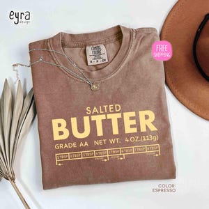 May include: Espresso-colored t-shirt with the words "SALTED BUTTER" in large yellow letters. The shirt also features a measurement guide and the text "NET WT. 4 OZ. (113g)". A silver necklace and a brown hat are also visible.