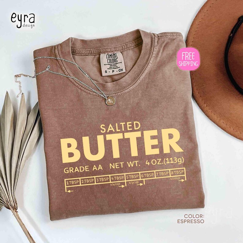 May include: Espresso-colored t-shirt with the words "SALTED BUTTER" in large yellow letters. The shirt also features a measurement guide and the text "NET WT. 4 OZ. (113g)". A silver necklace and a brown hat are also visible.