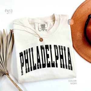 May include: An ivory short-sleeved t-shirt with a crew neckline and a Comfort Colors label. The shirt features the word PHILADELPHIA printed in a large, distressed black arched font across the chest. A thin gold chain necklace rests on the fabric.
