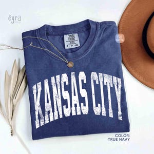 May include: A navy blue t-shirt with the text "KANSAS CITY" in distressed white lettering. The shirt has a crew neck and a small tag. A gold necklace with a pendant is draped across the shirt. The color is true navy.