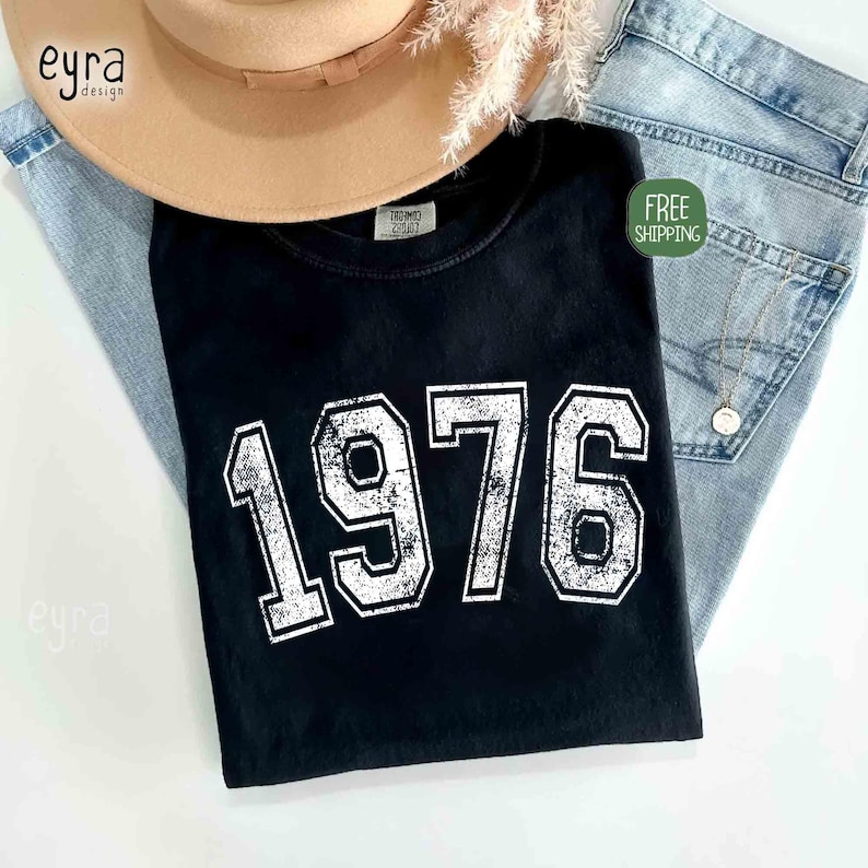 May include: Black t-shirt with the year "1976" printed in white, distressed lettering. The shirt is displayed with a tan hat and light blue jeans. A small green sticker with the words "FREE SHIPPING" is visible.