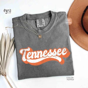 May include: A grey t-shirt featuring the word "Tennessee" in white with an orange outline. The shirt is folded and has a small tag at the neckline. A gold necklace and a brown hat are also visible.