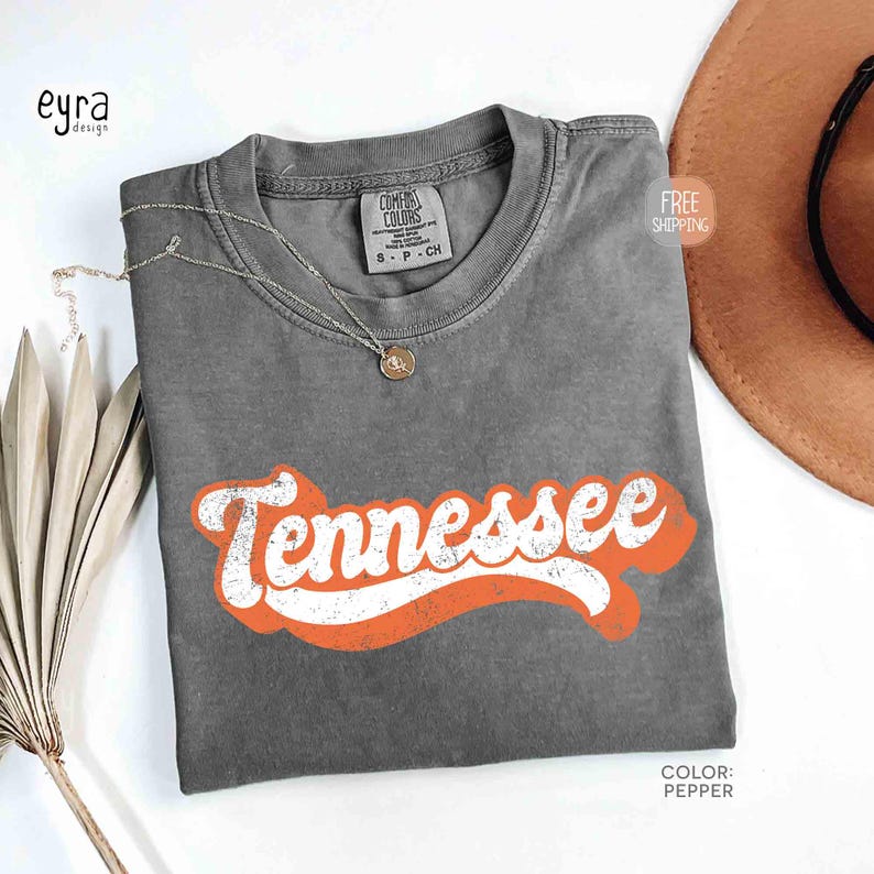 May include: A grey t-shirt featuring the word "Tennessee" in white with an orange outline. The shirt is folded and has a small tag at the neckline. A gold necklace and a brown hat are also visible.