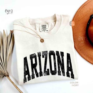 May include: An ivory short-sleeved t-shirt with a crew neckline featuring the word ARIZONA printed in a distressed black font across the chest. The shirt has a Comfort Colors label at the neck and is paired with a gold-toned pendant necklace.