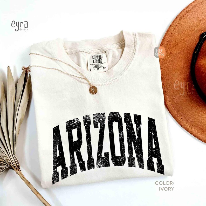 May include: An ivory short-sleeved t-shirt with a crew neckline featuring the word ARIZONA printed in a distressed black font across the chest. The shirt has a Comfort Colors label at the neck and is paired with a gold-toned pendant necklace.