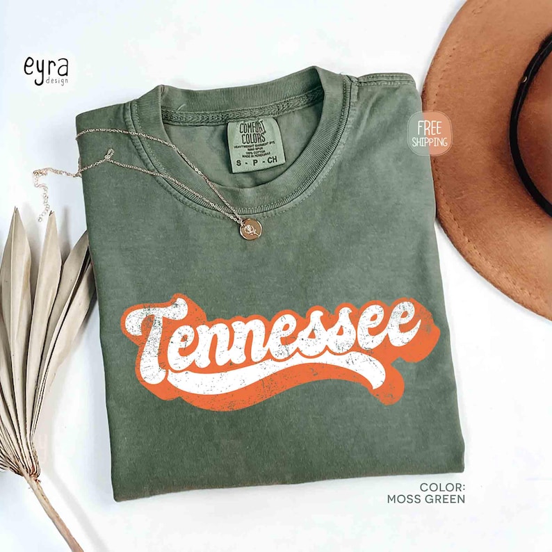 May include: A moss green t-shirt featuring the word "Tennessee" in white with an orange outline. A gold necklace with a pendant is on the shirt. The shirt is part of a clothing collection.