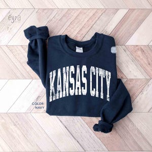 May include: A navy blue sweatshirt featuring the white text "KANSAS CITY" in an arched design. The garment is laid on a light brown and white wooden surface. The colour of the sweatshirt is labelled as "NAVY".