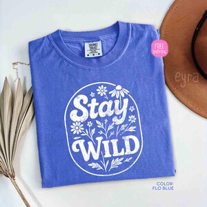 May include: A Flo Blue t-shirt with a white floral design and the words "Stay WILD" in a circular design. The shirt is a medium size and has a tag at the top. A brown hat is in the background.