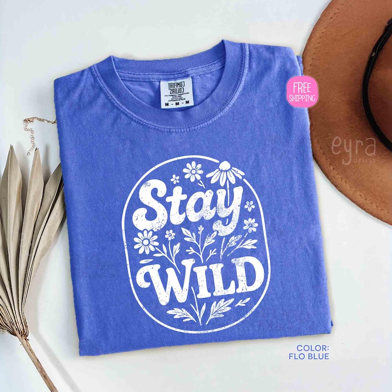 May include: A Flo Blue t-shirt with a white floral design and the words "Stay WILD" in a circular design. The shirt is a medium size and has a tag at the top. A brown hat is in the background.