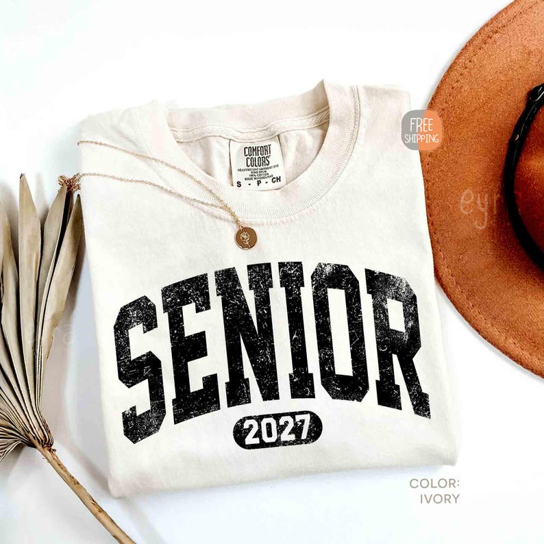 May include: Ivory-colored Comfort Colors t-shirt with the word "SENIOR" in distressed black lettering and the year "2027" below. A gold necklace with a pendant rests on the shirt. A brown hat is partially visible in the background.