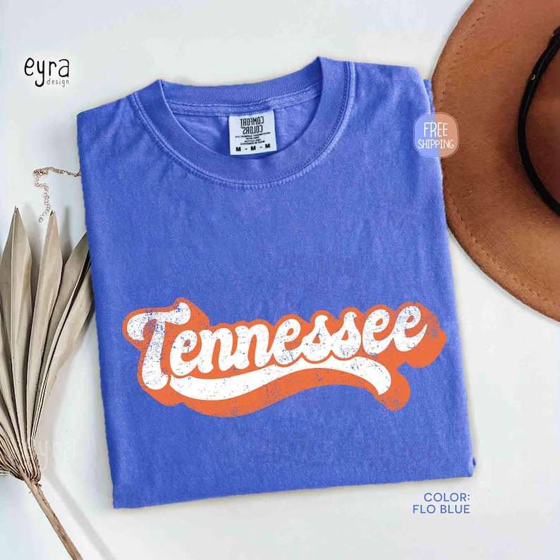 May include: A blue t-shirt featuring the word "Tennessee" in white with an orange outline. The shirt is folded and has a label at the neckline. The colour of the shirt is "Flo Blue".