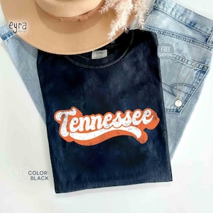 May include: A black t-shirt featuring the word "Tennessee" in white and orange. The shirt is displayed with a tan hat and light blue jeans. The text "Color: Black" is visible.