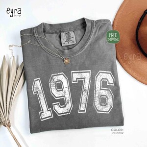 May include: A gray Comfort Colors t-shirt with the year "1976" printed in white, featuring a distressed, vintage style. A gold necklace rests on the shirt. The shirt is labeled "Pepper" in color.