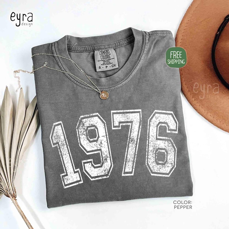 May include: A gray Comfort Colors t-shirt with the year "1976" printed in white, featuring a distressed, vintage style. A gold necklace rests on the shirt. The shirt is labeled "Pepper" in color.